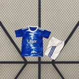 Equipación Como 25-26 local para niño vista frontal con camiseta azul de gráfico blanco y pantalón corto blanco