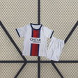 Equipación PSG 26-27 visitante niño vista frontal con camiseta blanca, franja central azul marino y roja y pantalón blanco