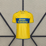Vista frontal centrada de la camiseta Las Palmas 26-27 local en amarillo, con banda azul horizontal, cuello redondo y detalles azules
