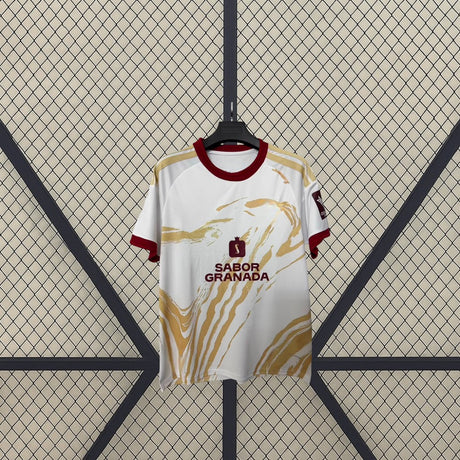 Vista frontal de la camiseta Granada 26-27 tercera equipación en blanco con gráfico dorado y cuello redondo granate