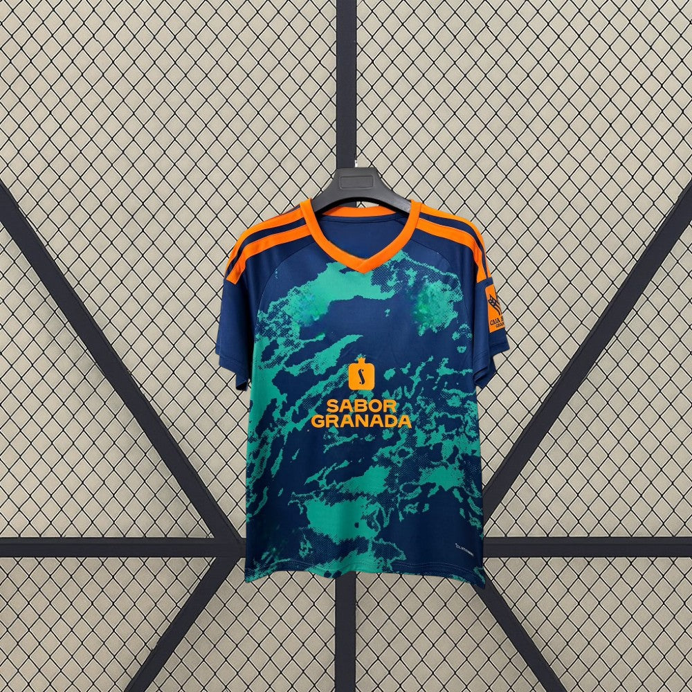 Vista frontal de la camiseta Granada 26-27 visitante en azul marino con gráfico verde, cuello en V naranja y detalles naranjas en hombros