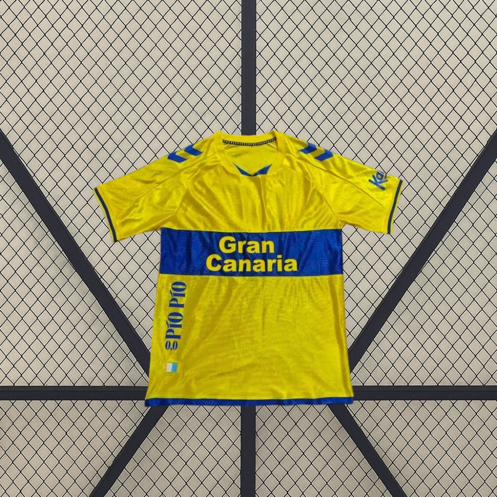 Vista frontal de la camiseta Las Palmas 26-27 local extendida, con detalles azules en hombros, puños y bajo y textura lineal