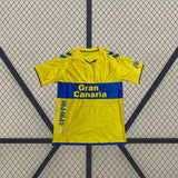 Vista frontal de la camiseta Las Palmas 26-27 local extendida, con detalles azules en hombros, puños y bajo y textura lineal