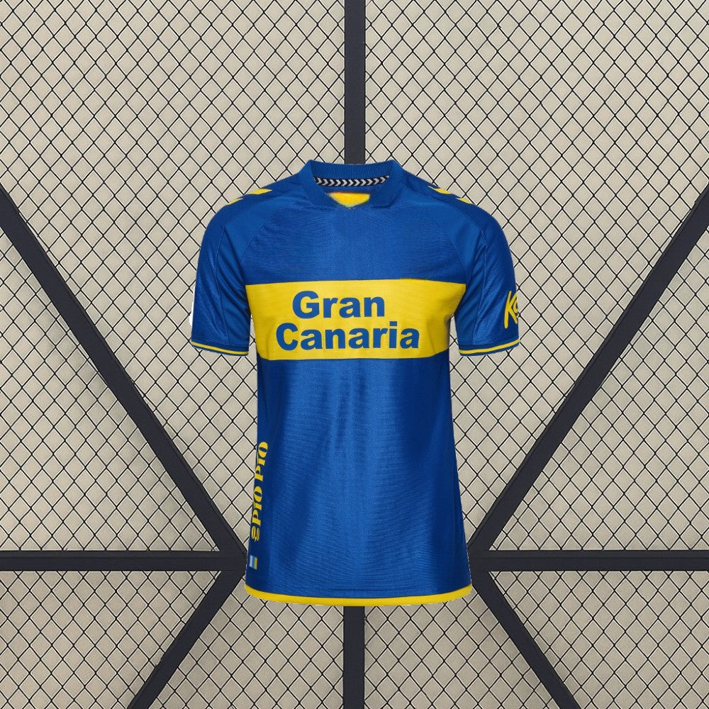 Vista frontal de la camiseta Las Palmas 26-27 visitante en azul, con banda amarilla horizontal, cuello redondo y manga corta