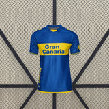 Vista frontal de la camiseta Las Palmas 26-27 visitante en azul, con banda amarilla horizontal, cuello redondo y manga corta