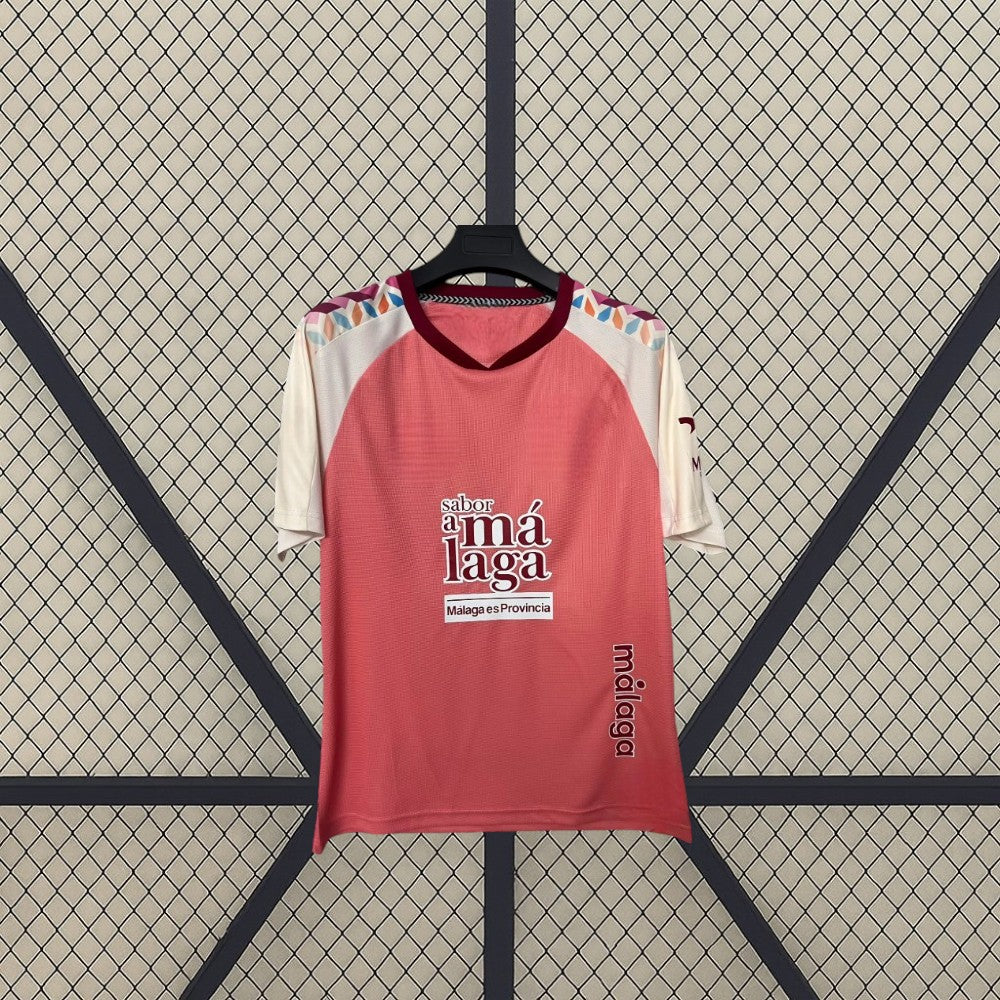 Vista frontal de la camiseta Málaga 26-27 visitante colgada, con base coral, mangas crema y detalles geométricos multicolor en los hombros