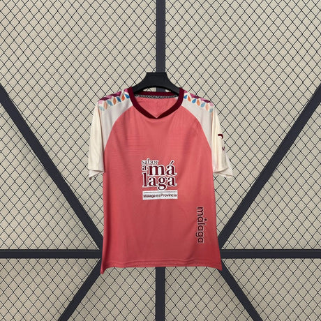 Vista frontal de la camiseta Málaga 26-27 visitante colgada, con base coral, mangas crema y detalles geométricos multicolor en los hombros