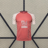 Vista frontal de la camiseta Málaga 26-27 visitante en color coral, con mangas crema, cuello oscuro y textura vertical en el tejido