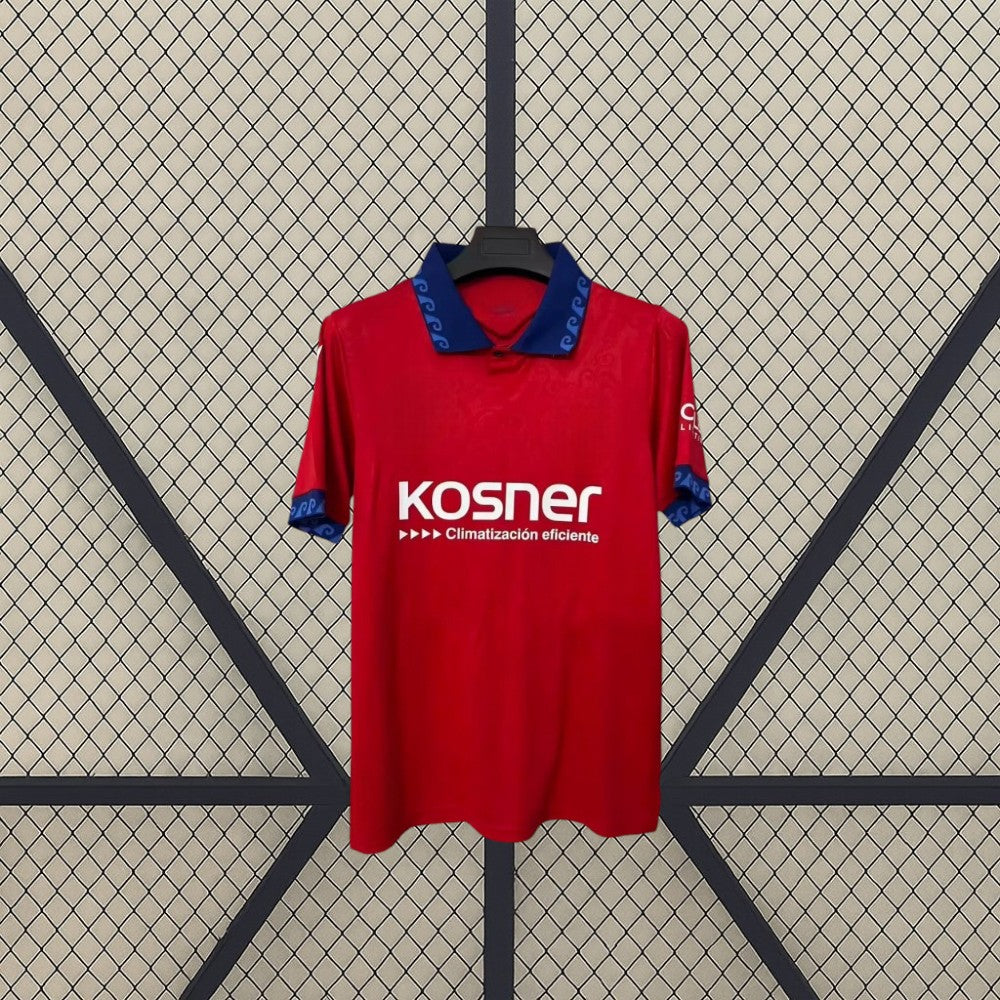 Vista frontal de la camiseta Osasuna 26-27 local colgada, con base roja, cuello azul y detalles gráficos en cuello, pecho y mangas