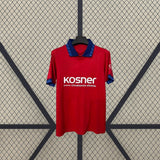 Vista frontal de la camiseta Osasuna 26-27 local colgada, con base roja, cuello azul y detalles gráficos en cuello, pecho y mangas