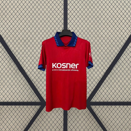 Vista frontal de la camiseta Osasuna 26-27 local colgada, con base roja, cuello azul y detalles gráficos en cuello, pecho y mangas