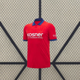 Vista frontal de la camiseta Osasuna 26-27 local en rojo, con cuello tipo polo azul, puños estampados y patrón tonal en el pecho