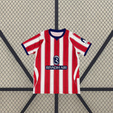 Vista frontal extendida de la camiseta Atlético de Madrid 26-27 local, con rayas rojas y blancas, detalles azules y paneles laterales claros