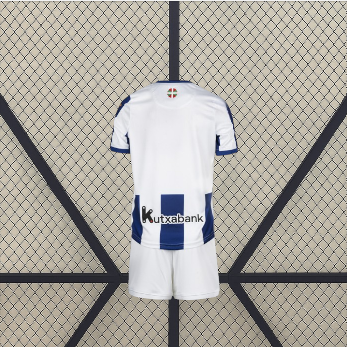 Vista trasera de la equipación Real Sociedad 26-27 local para niño con espalda blanca, detalle de franjas azules en la parte inferior y pantalón blanco