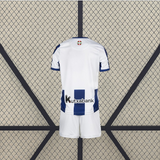 Vista trasera de la equipación Real Sociedad 26-27 local para niño con espalda blanca, detalle de franjas azules en la parte inferior y pantalón blanco